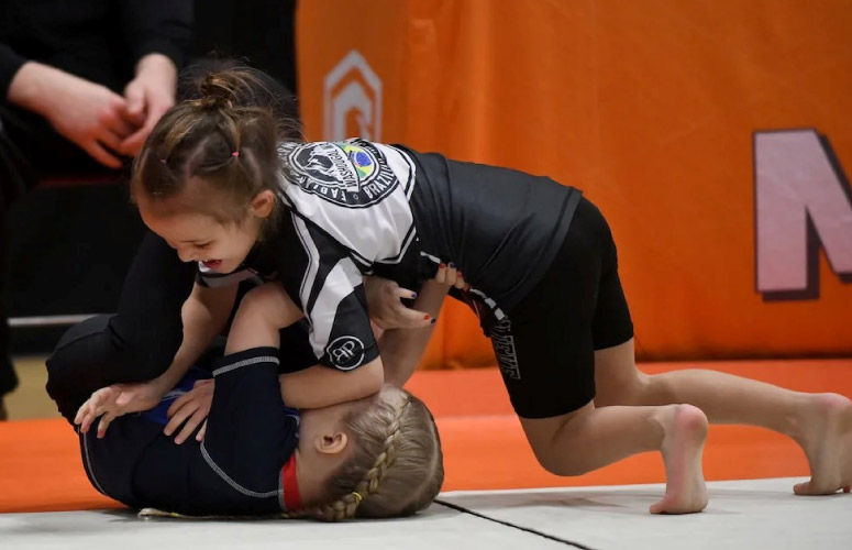 Two young Caucasian girls grapple at a tournament. One girl has her back on the mat trying to recover guard and the other girl tries to establish side control. The girl on bottom has blonde hair in two braids and the girl on top has brunette hair in a roped pony tail. Both girls are wearing black rash guards and leggings.