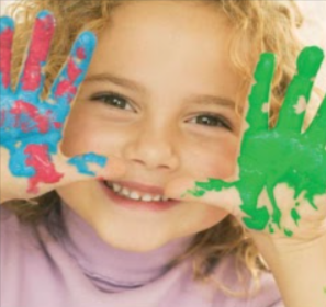A happy little girl with a huge smile poses showing her paint covered hands to the viewer. She has shoulder length curly blond hair. The paint on her hands is green, pink, and blue.