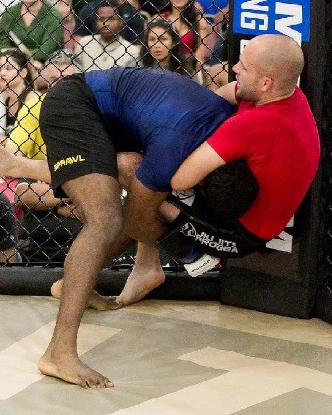 Don Stoner holds a flying guillotine on an opponent in a cage grappling match. He is wearing a red rash guard and black shorts. His opponent, who's face cannot be seen, is wearing a blue rash guard and black shorts.