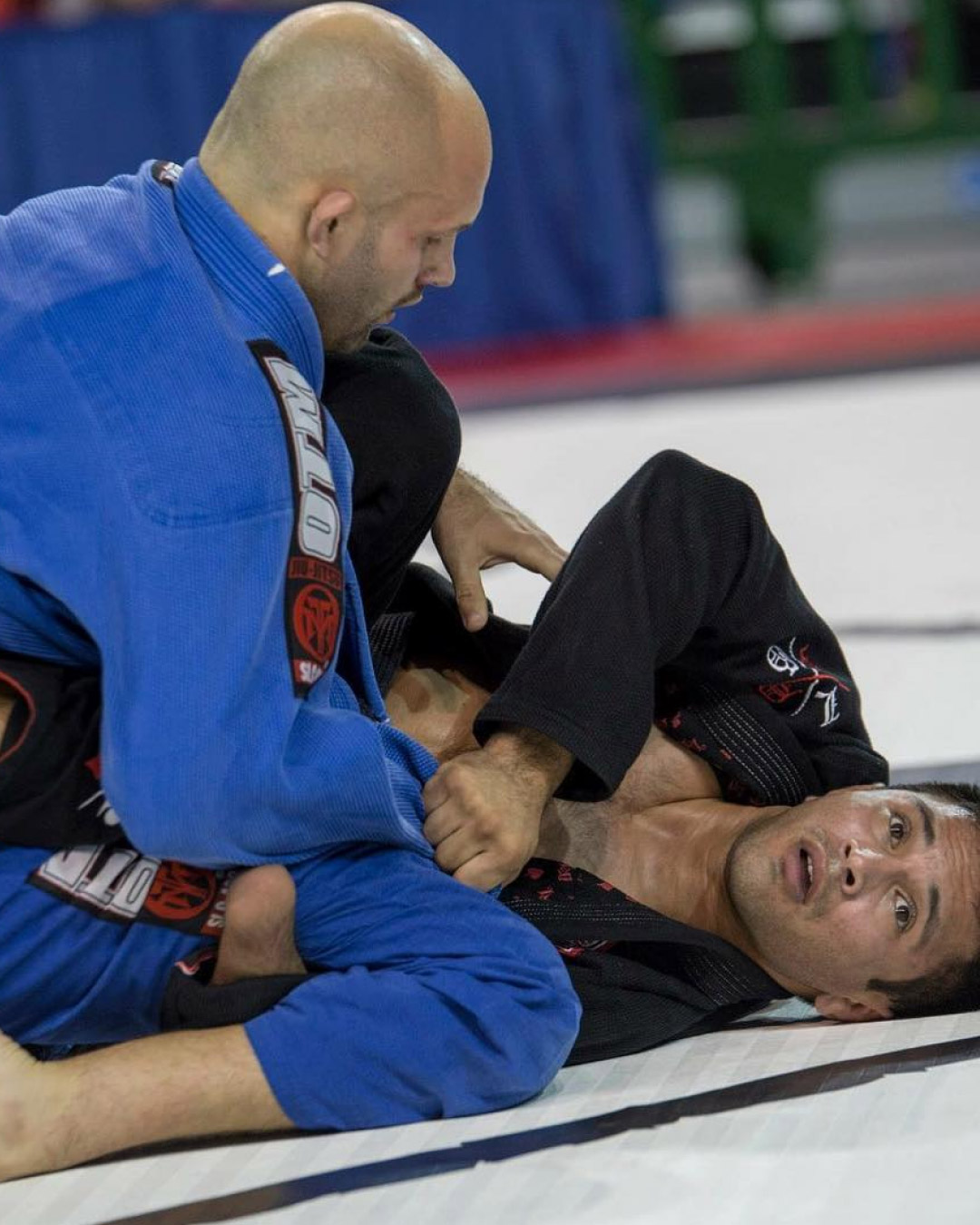 Two men grapple at a tournament. The man on bottom tries to recover guard while looking to his coach for support. He is wearing a black martial arts gi. The man on top works to establish a dominant position. He is wearing a blue gi.