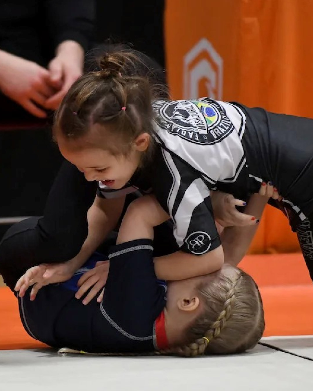 Two young Caucasian girls grapple at a tournament. One girl has her back on the mat trying to recover guard and the other girl tries to establish side control. The girl on bottom has blonde hair in two braids and the girl on top has brunette hair in a roped pony tail. Both girls are wearing black rash guards and leggings.