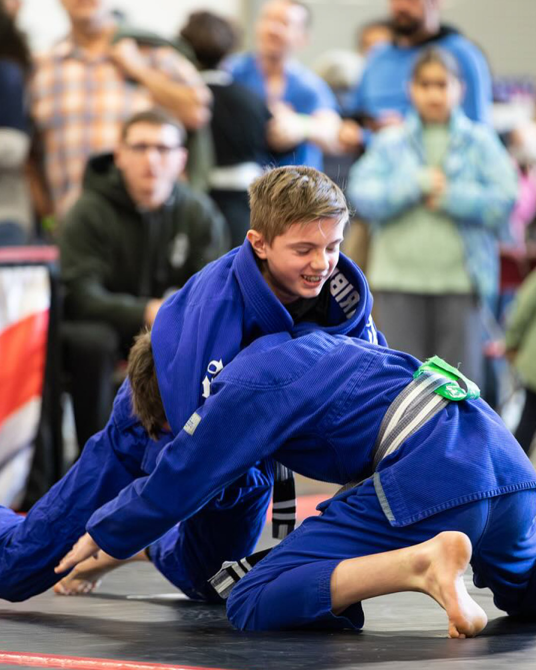 Two boy grapple at a jiu-jitsu tournament. The boy on top is smiling and has blond hair. He is holding the start of a guillotine on the other boy who is on his knees attempting to grab the knees of his competitor. Both are wearing blue martial arts gis. A coach can be seen in the background wearing glasses and black pants with a black hoodie. There is a crowd of onlookers.