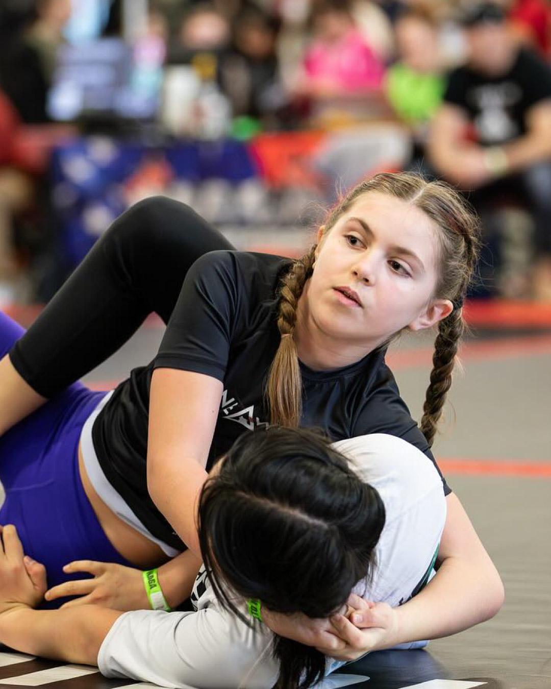 Two girls grapple at a jiu-jitsu tournament. One girl is on her back and is wearing a white rash guard and black leggings. She has black hair and is trying to recover guard. The girl on top is in side control and is wearing a black Lone Wolf Jiu-Jitsu rash guard and blue leggings. She has blonde hair in two braids and is looking to her coach for instructions.