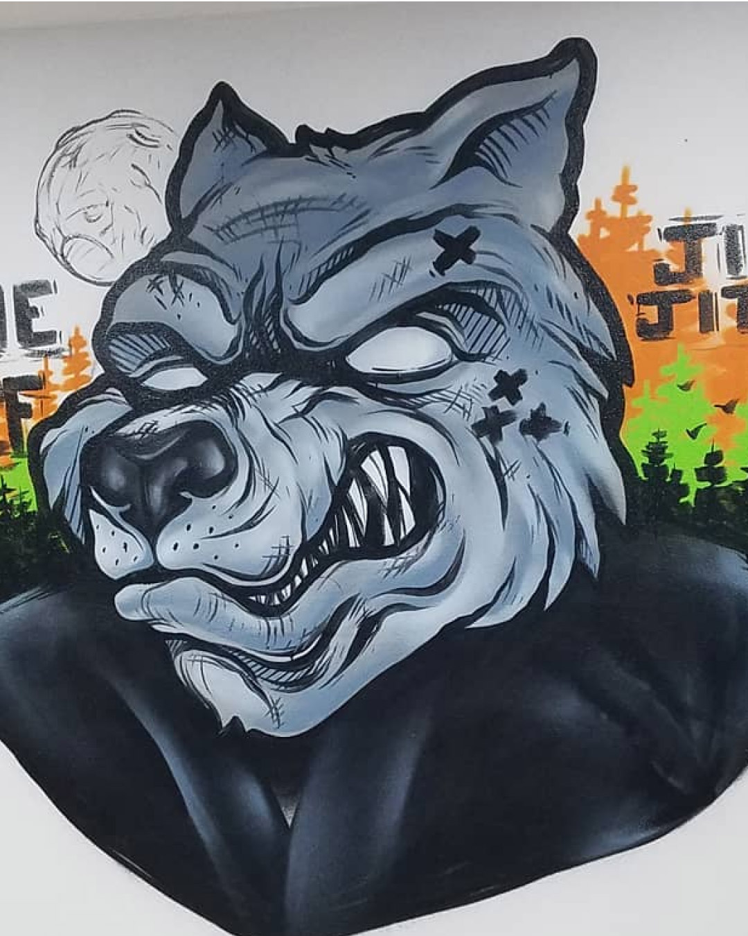 A painting of a serious gray wolf on a white wall with the words "Lone Wolf Jiu-Jitsu". The wolf wears a black martial arts gi. There is a moon in the distance with green and orange colors.