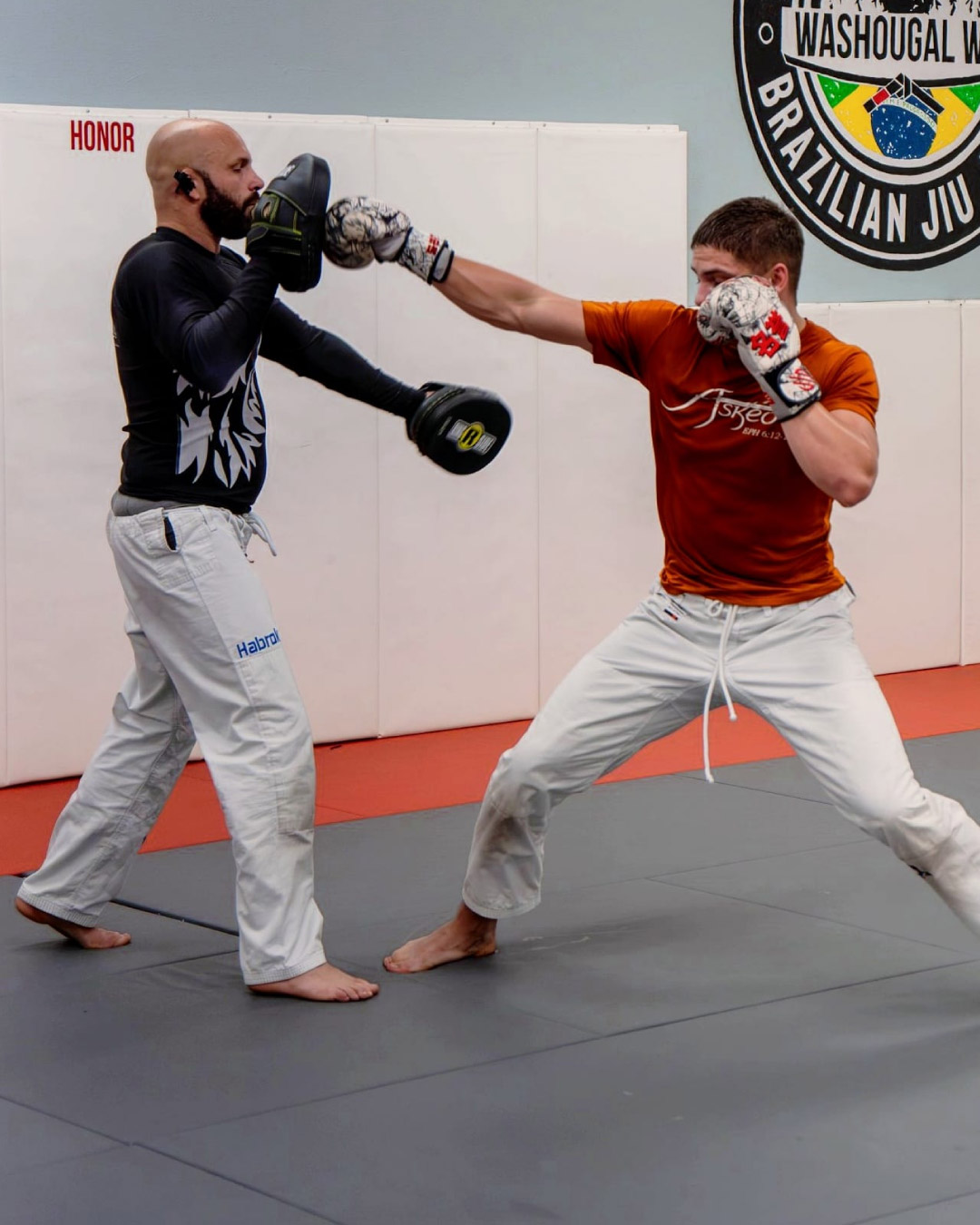 A Caucasian male with a red shir and white gloves practices throwing an straight punch with a partner. His hair is very short and brown. His partner is bald with a beard and goatee and is wearing a black rash guard with punching mitts.