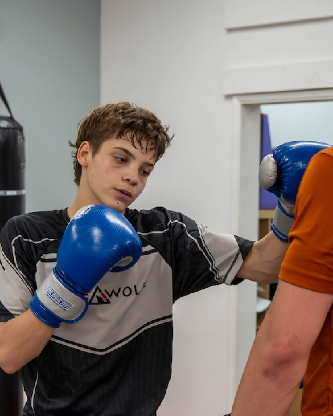 A Caucasian short-haired blond teen performs a punch on an off screen student. The teen is wearing blue gloves and a black and white striped shirt with the Lone Wolf Jiu-Jitsu logo on it. A black heavy bag is in the background.