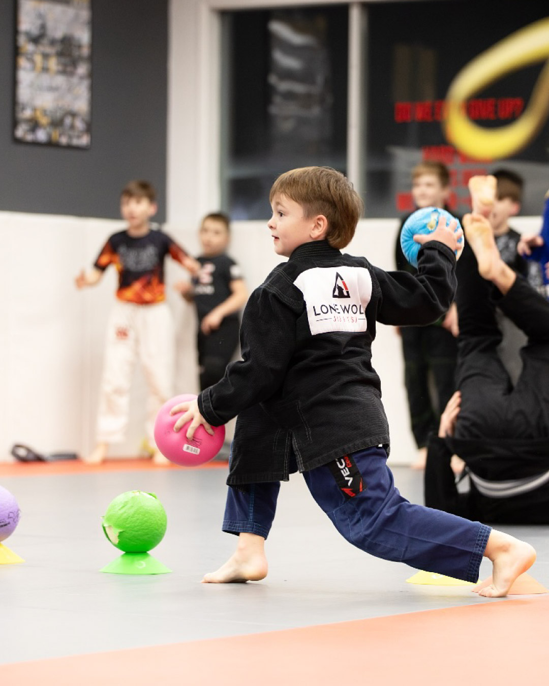 A young male Caucasian child in a strong lunging pose holds a pink dodge ball while attempting to throw a blue one with his other hand. He is wearing a black gi top with the Lone Wolf Jiu-Jitsu logo on it with dark blue gi panes. There are more dodge balls lined up in front of him. Behind him are other students who have been knocked out of the game.