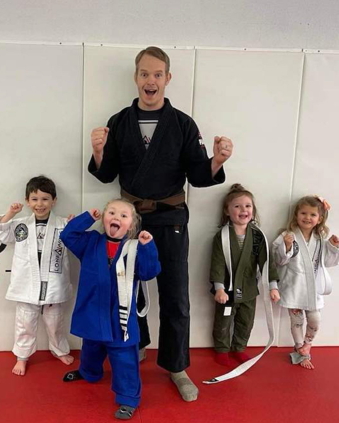 A martial arts instructor poses with his ninja club students. Everyone makes funny faces after class. The teacher is wearing a black martial arts gi while the children wear either white, blue, or grey uniforms.