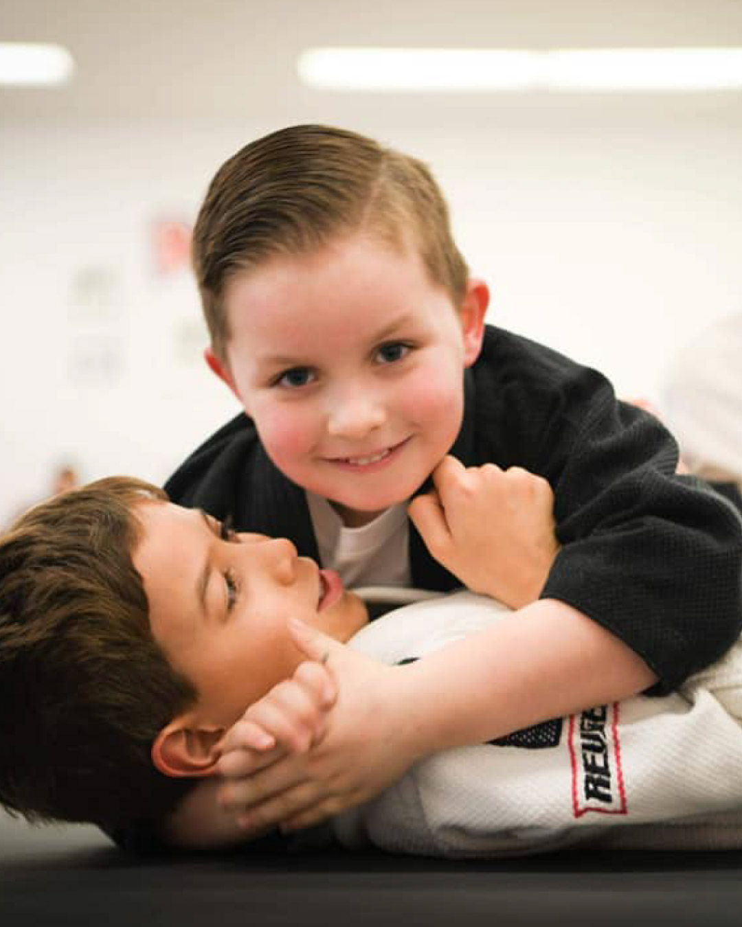 Two very young children wrestle on the ground. One on the ground is laying on his back and has short brown hair and and a white martial arts gi. The other child is blond and is laying on him perpendicular in side control and is wearing a black martial arts gi.
