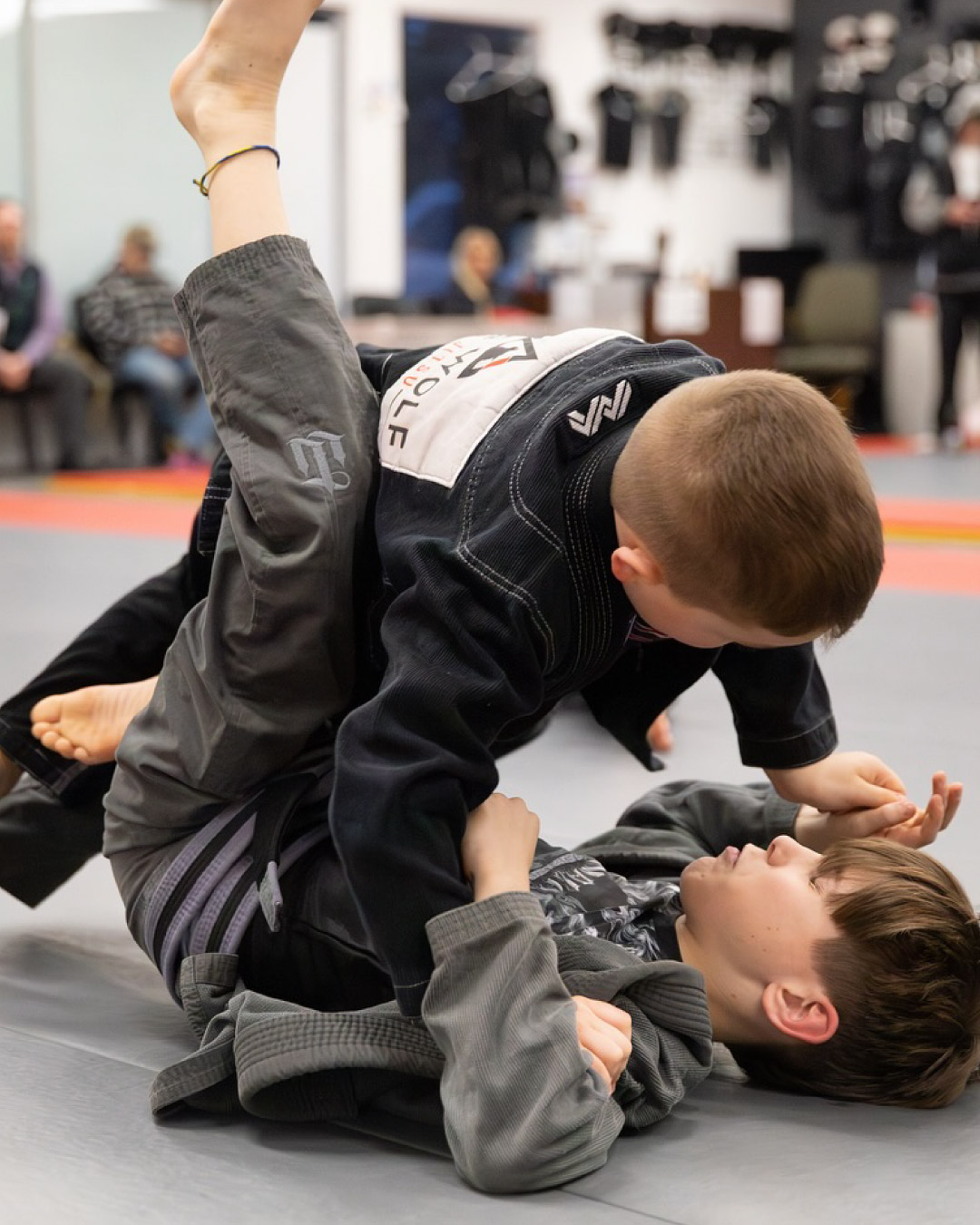 Two Caucasian males students grapple on black and red mats. Bot children has short hair and black martial arts gis with Lone Wolf Jiu-Jitsu logos on them. One child is laying on his back and the other child is trying to pass his guard.