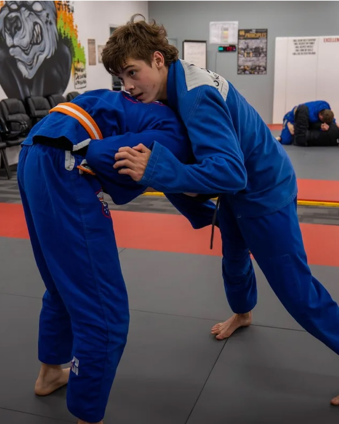 Two students wearing blue martial arts gis with Lone Wolf Jiu-Jitsu logos grapple while standing up on black mats. A wolf painting is on the wall in the background on the left and a mirror reflects the scene on the right.