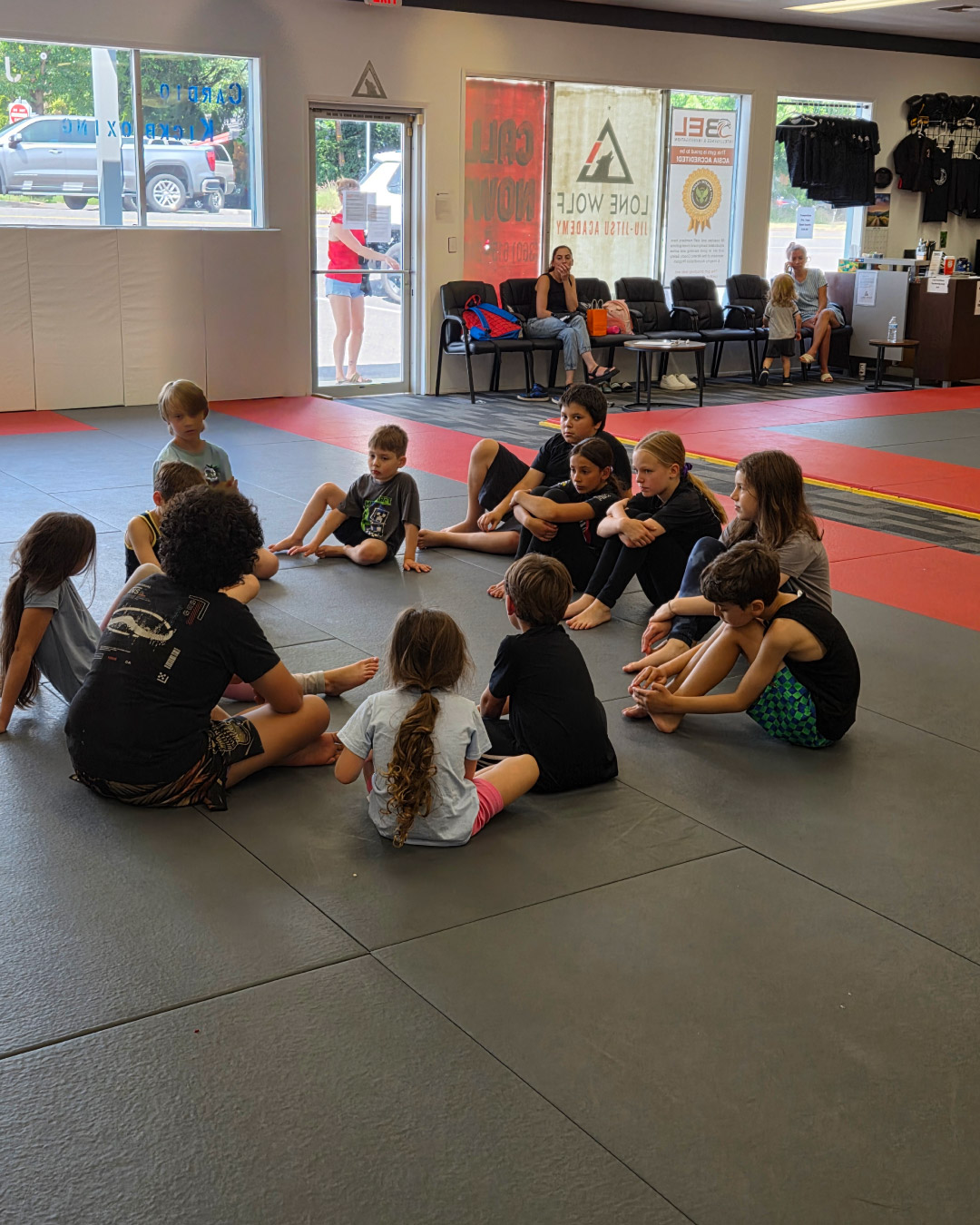 A group of students sit on a black and red mats talking to each other about jiu-jitsu. The students are young, teen and under age group, and of various genders and races. Parents sit on black chairs watching in the background. The Lone Wolf Jiu-Jitsu logo can be seen on the window in the background.