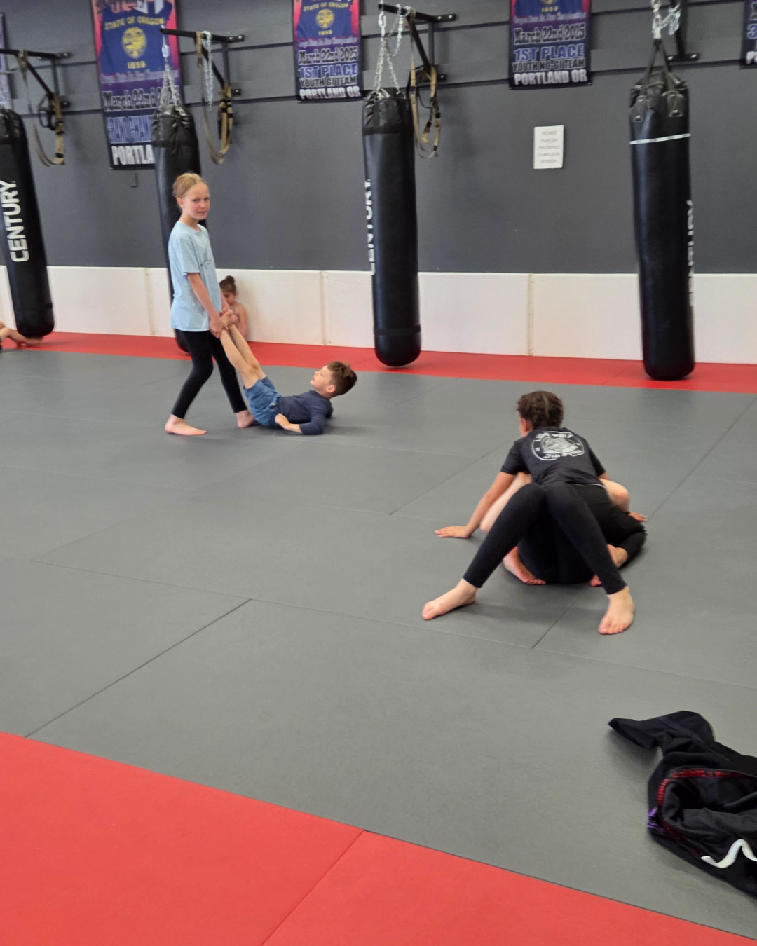 A group of children practice martial arts at Lone Wolf Jiu-Jitsu. The children are various ages, races, and genders. The mats are black and red. There are black heavy bags in the background.