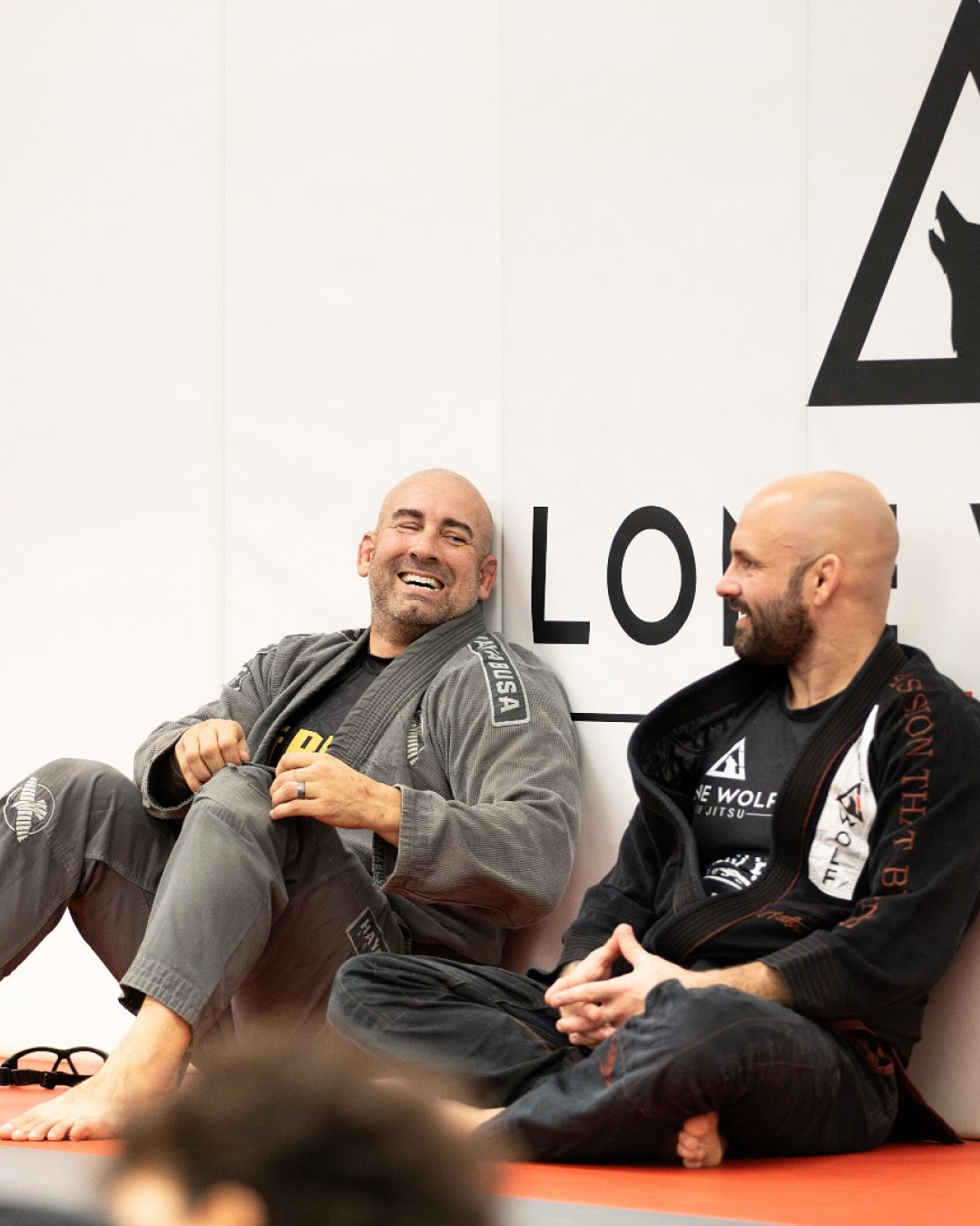 Two men take a break during Brazilian Jiu-Jitsu class at Lone Wolf Jiu-Jitsu. They are smiling and laughing. One wears a gray martial arts uniform and the other wears a black uniform.