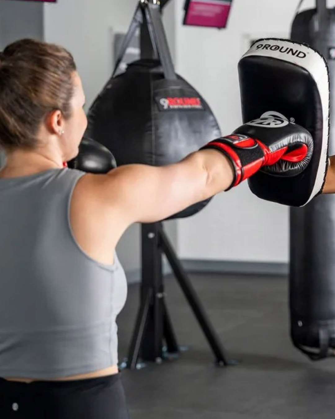 A woman, facing away from the camera, strikes a punching mitt. She is wearing a gray tank top. There are heavy bags in the background.