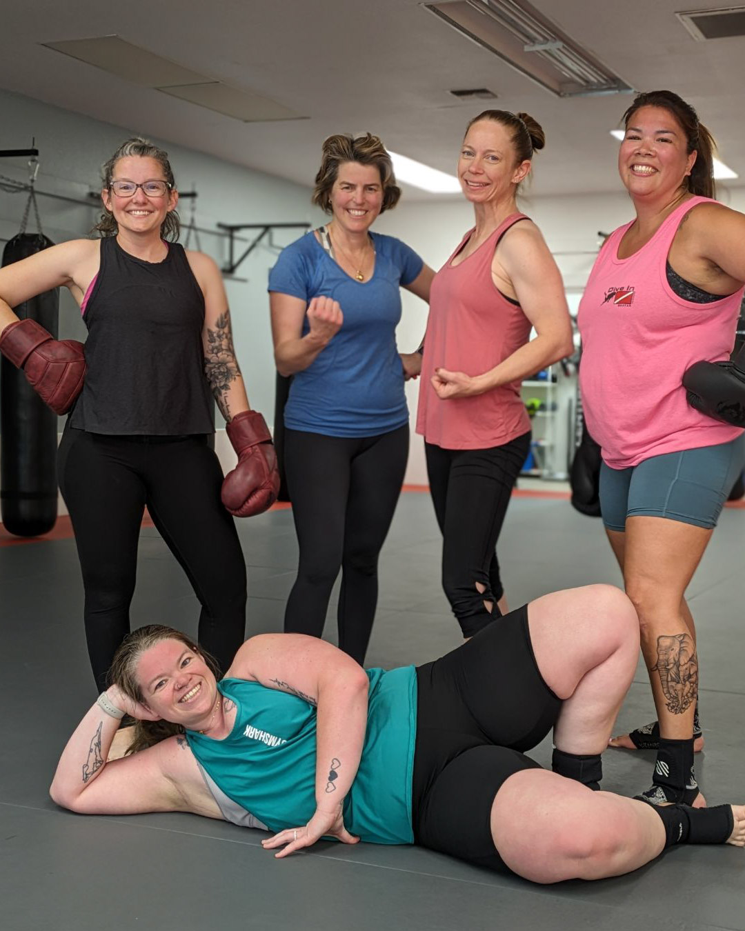 A group of five women pose at Lone Wolf Jiu-Jitsu after an intense fitness kickboxing class. One woman lays sprawled out on the ground wearing a turquoise tank top and black short leggings. The other women are standing in various poses wearing either black leggings or grey legging shorts. The two on the right have pink tank tops. The woman on the far left has glasses and a black tank top. The second to the left has a blue t-shirt.