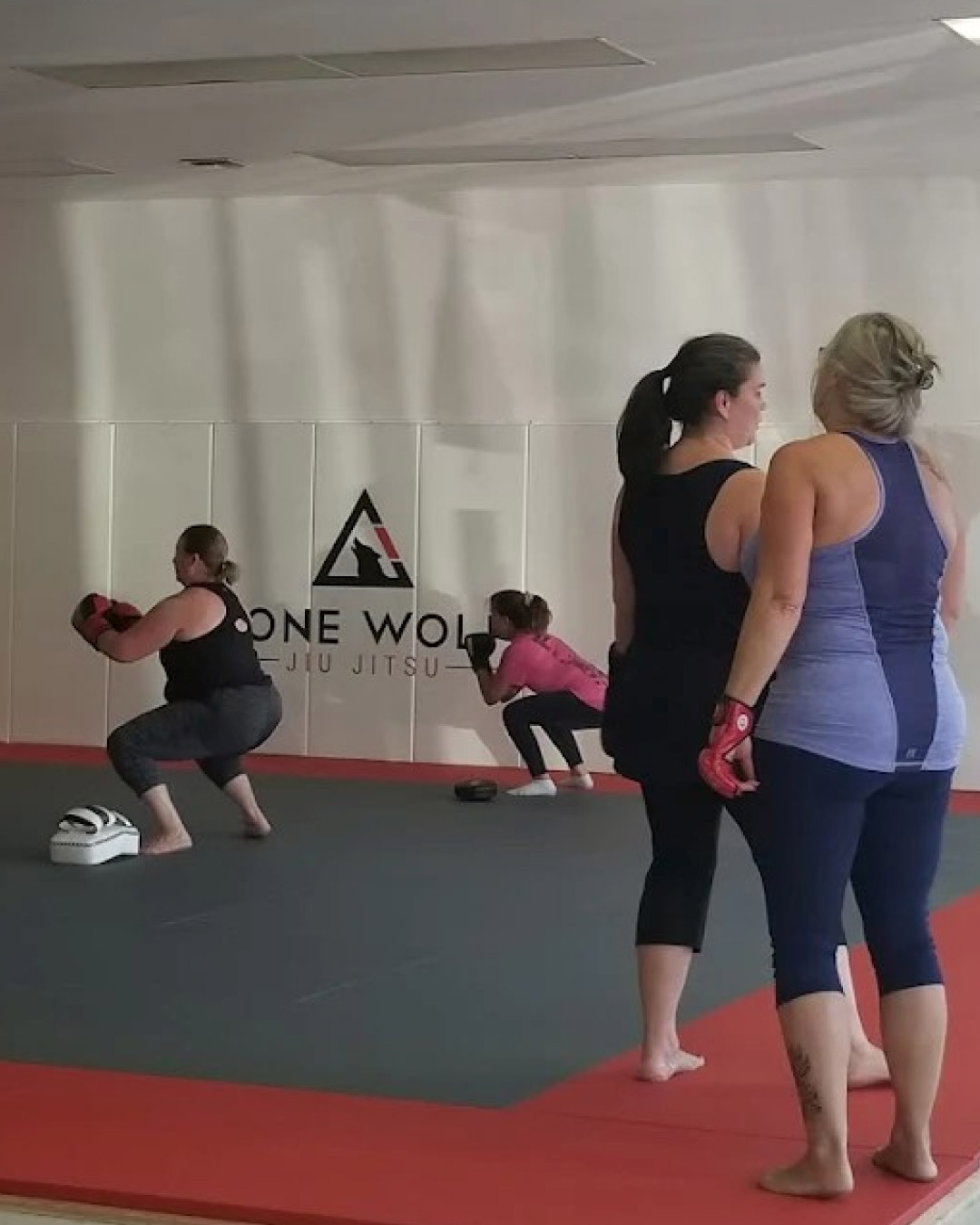 A group of women practice fitness kickboxing squats in front of a white wall with a Lone Wolf Jiu-Jitsu logo. Two women are standing taking a break in the foreground. The mats are black and red.