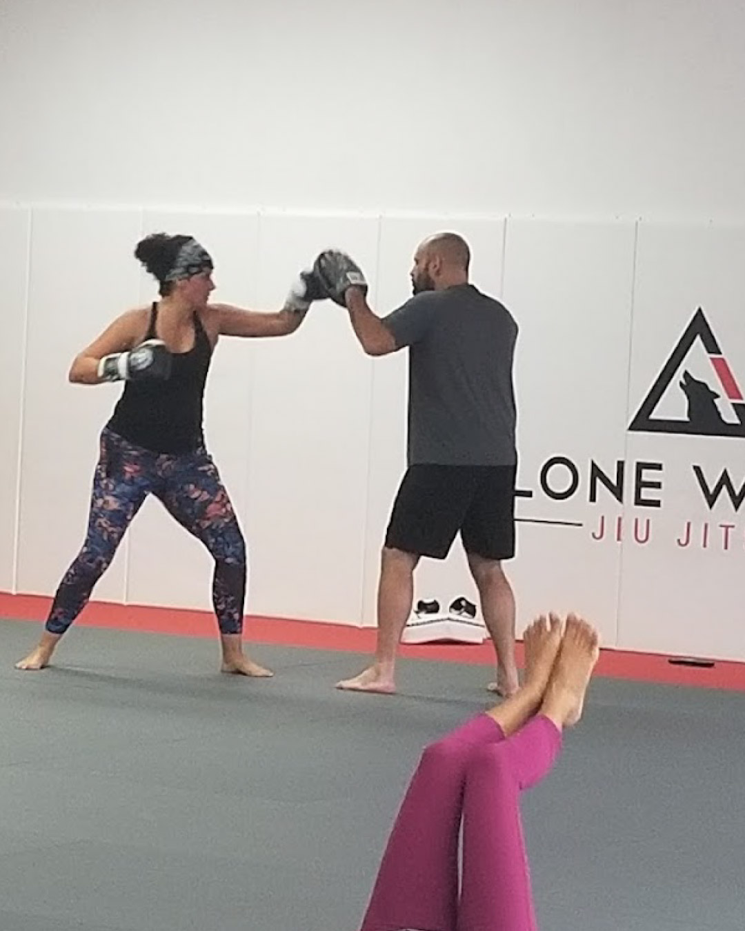 A brunette women woman wearing a black tank top and camo leggings practice fitness kickboxing by throwing a straight punch at a black mixed race male wearing a grey t-shirt and black shorts in front of a white wall with the Lone Wolf Jiu-Jitsu logo. A woman in the foreground wearing pink leggings does leg lifts.