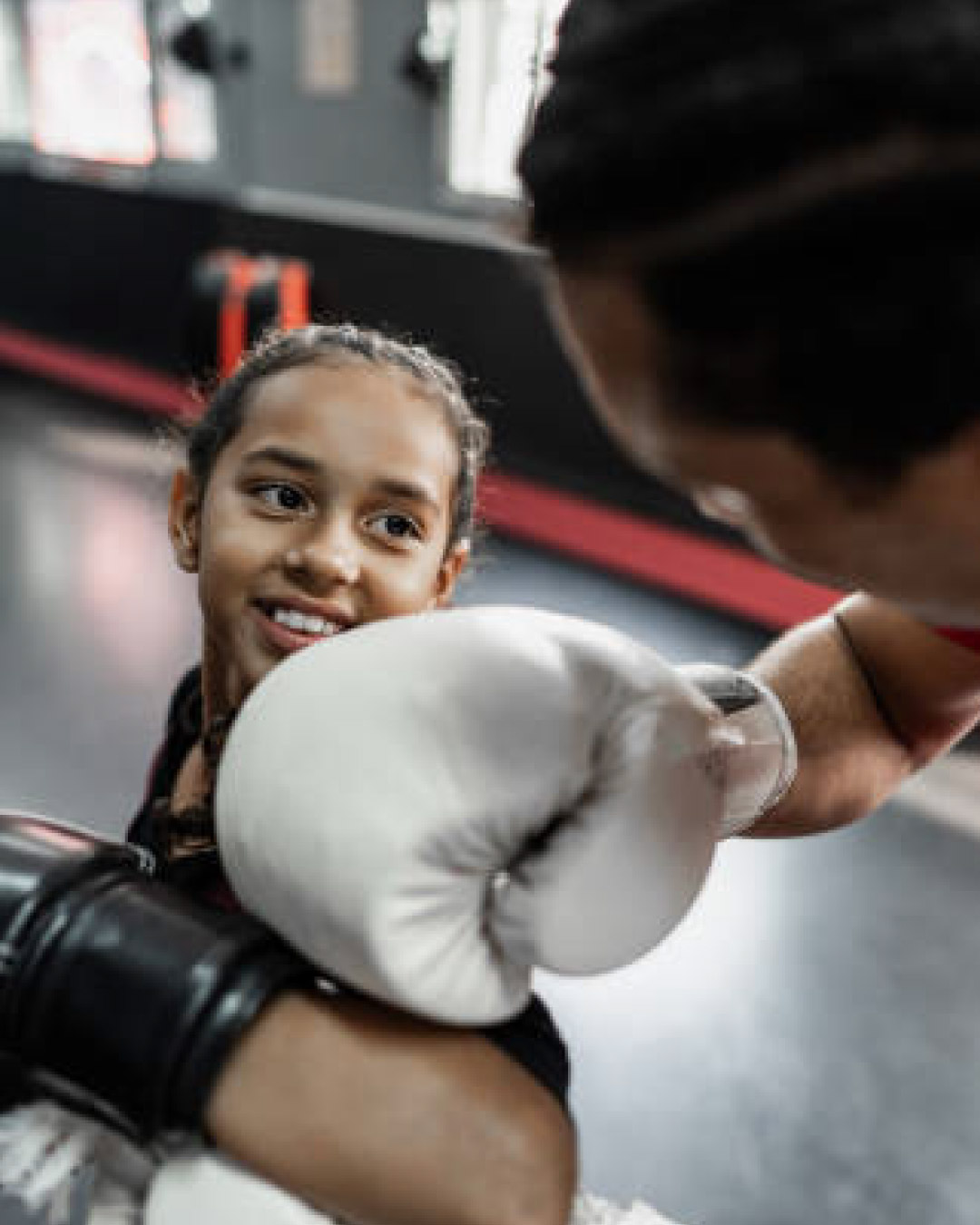 A closeup shot of a girl of unknown race and ethnicity blocking a punch from an instructor. She is smiling and is wearing black gloves and a black t-shirt. The gym mats behind her are black.