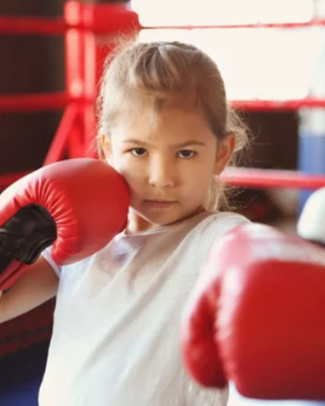A blonde little girl strikes a martial arts pose with a serious face inside of a boxing ring. She has blonde hair, red gloves, and a white t-shirt.
