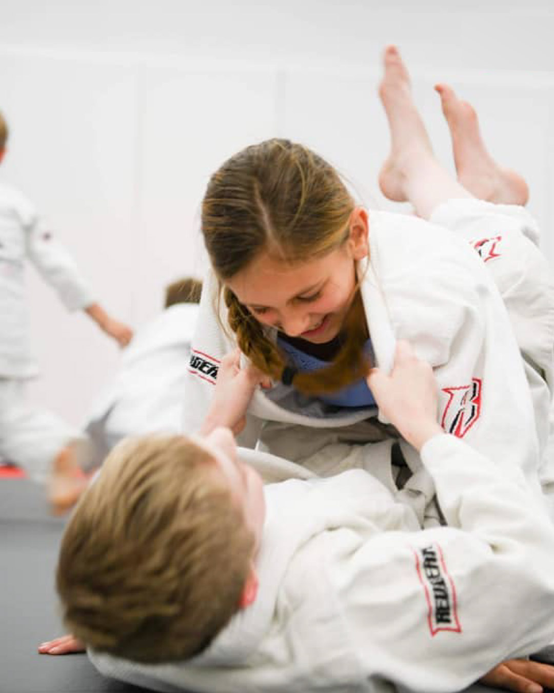 Two Caucasian students practice Brazilian Jiu-Jitsu. One boy is laying on his back while the other, a girl, tries to pass his guard. The boy on his back has short blonde hair and the girl has blonde hair in a braid. Both wear white martial art gis. The mats are black and red. The background is white.