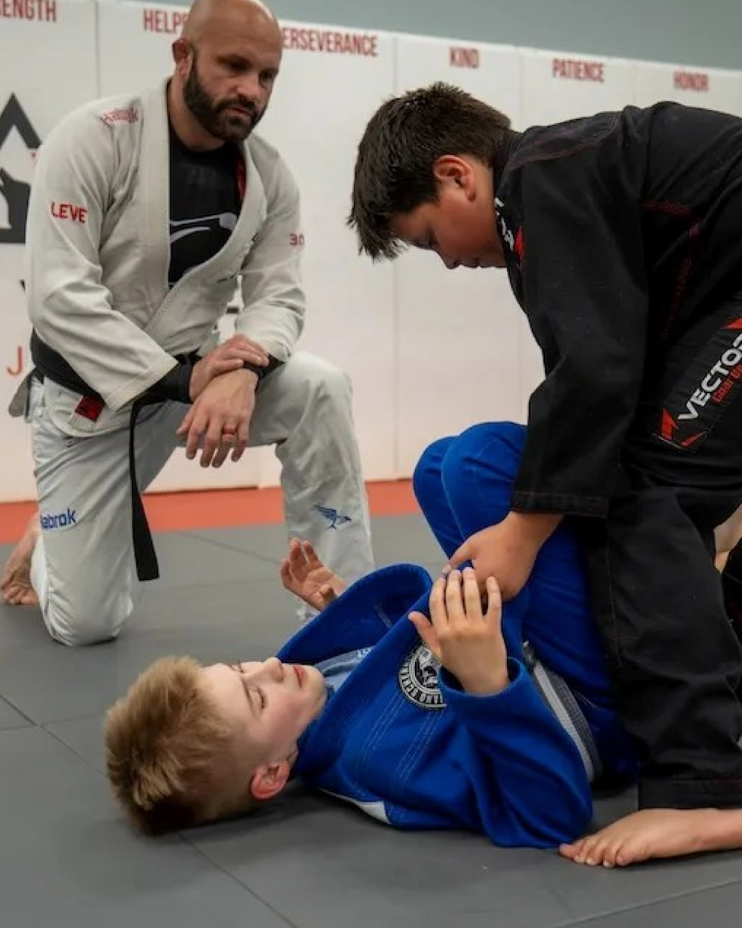 A bald, Caucasian man with a short black beard and goatee wearing a white martial arts gi teaches two Caucasian, children Brazilian Jiu-Jitsu. One child is laying on the ground with his short, blonde hair wearing a blue gi and the other is standing over him trying to pass his guard. He is wearing a black martial arts gi and has short, black hair. A white wall in the background says "Kind", "Patience", "Perseverance" and "Honor" with the Lone Wolf Jiu-Jitsu logo.