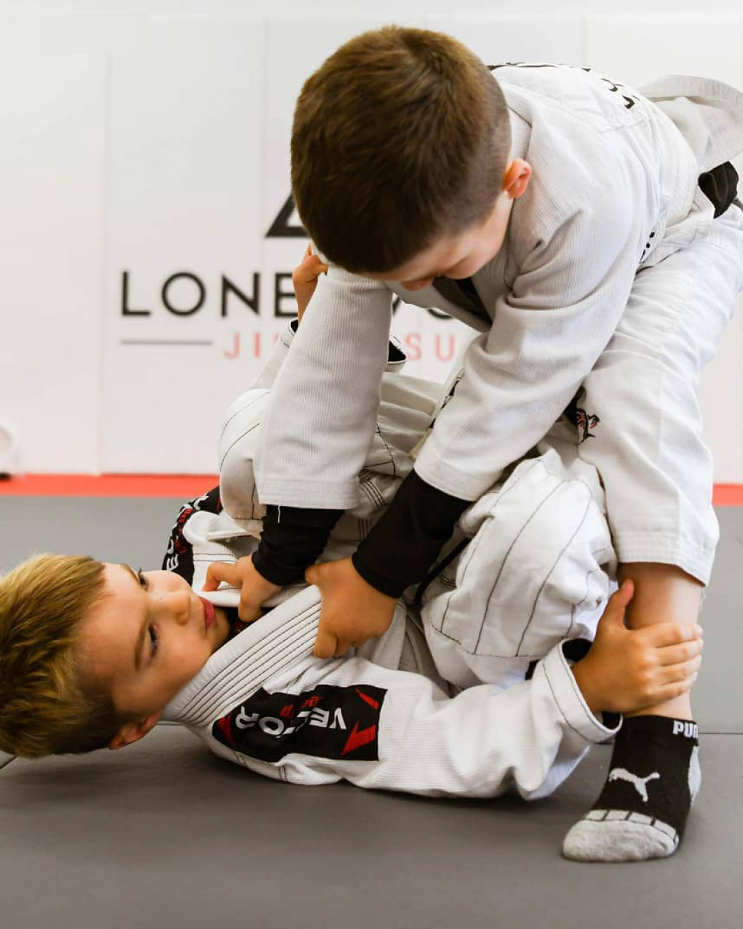Two Caucasian boys practice Brazilian Jiu-Jitsu. One boy is laying on his back while the other tries to pass his guard. The boy on his back has short blonde hair and the standing boy has brunette hair. Both wear white martial art gis. The mats are black and red. The background is white with a Lone Wolf Jiu-Jitsu logo.