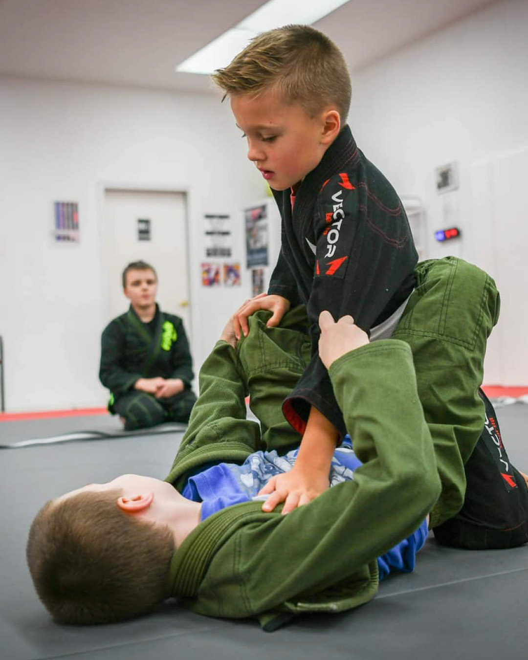 Two boys practice grappling moves while in class at Lone Wolf Jiu-Jitsu. The boy on top, wearing a black martial arts gi, is trying to pass guard while the boy on bottom, in a green martial arts gi, is trying an arm drag while looking for assistance from someone off screen. Another student watches in the background.