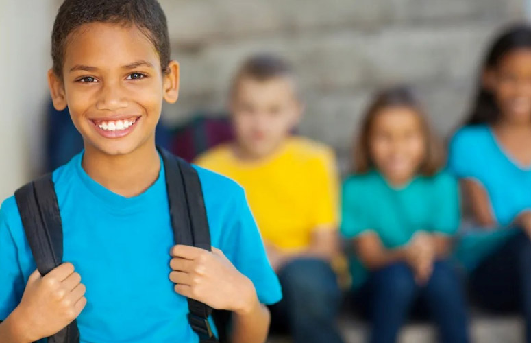 A child of unknown race smiles at the camera while wearing a blue shirt and backpack. There are other children in the background.