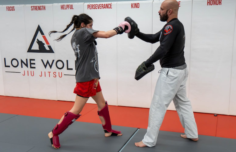 A female practices striking a mitt held by a male. She is wearing a gray t-shirt with red shorts along with pink boxing gloves and red shin guards. The man wears white martial arts gi pants with a black rash guard. They are standing on black and red mats in front of a white wall that say says "Focus", "Strength", "Perseverance", and "Helpful" with the Lone Wolf Jiu-Jitsu logo.