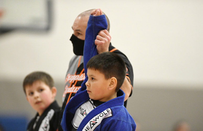 A male child has his hand raised after winning a grappling tournament. He wears a blue martial arts uniform with the Lone Wolf Jiu-Jitsu logo on it.