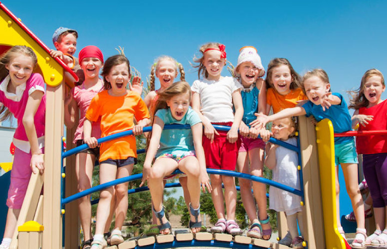 A group of children stand on a playground with happy faces. The children are of mixed races, ages, and genders.
