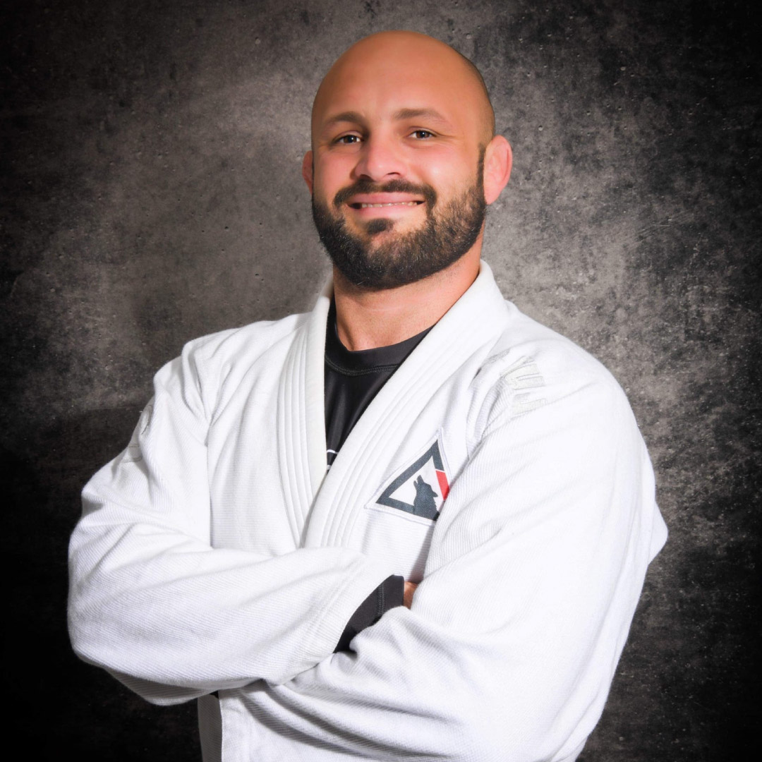 Head Coach Don Stoner poses for a photo in front of a standard black studio photography background. He is bald with a beard and goatee. He has a smile and a white martial arts gi with the Lone Wolf Jiu-Jitsu logo.