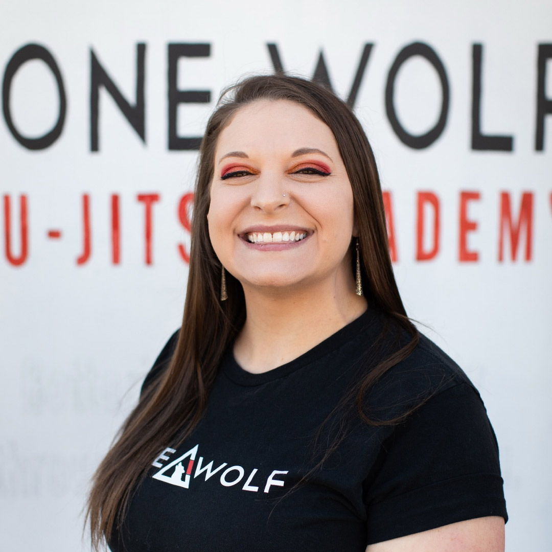 Robyn, the office assistant, poses in front of a white wall with the Lone Wolf Jiu-Jitsu Logo. She has a long hair style with make up, including red eye shadow and nude lips. Long earring dangle from her ears. She wears a black shirt with the Lone Wolf Jiu-Jitsu logo.