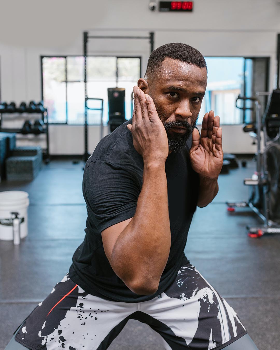 An African American male poses for the camera in a gym. He has his arms up by his face in a blocking manner. He is wearing a black shirt and black and white shorts.