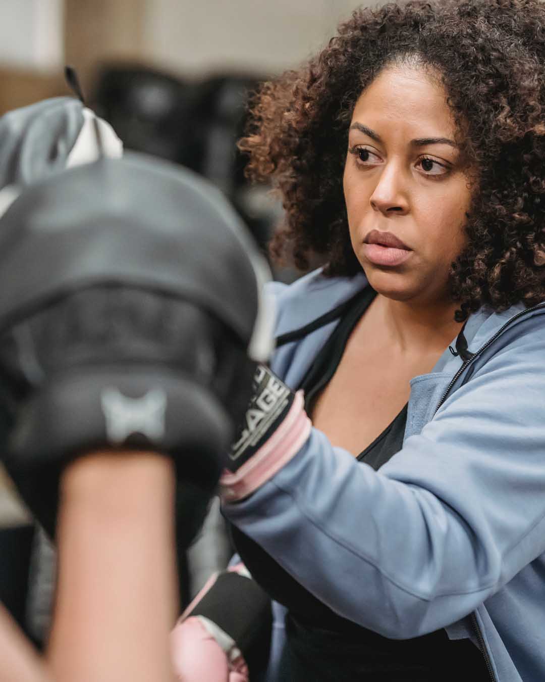 An African American woman with a traditional hair style practices fitness kickboxing by throwing punches and a set of mitts being help by a person who is off screen. The woman wears a black tank top with a blue zipper jacket over it.