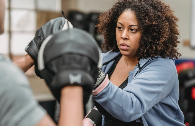 An African American woman with a traditional hair style practices fitness kickboxing by throwing punches and a set of mitts being help by a person who is off screen. The woman wears a black tank top with a blue zipper jacket over it.